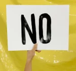 A person holding up a white sign with the word "NO" painted in bold black letters, against a yellow background.