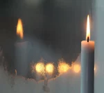 A lit candle in focus with a blurred reflection of its flame and three other candles on a frosted glass surface.