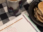 A close-up view of a ceramic mug filled with coffee, a plate with stacked pancakes, and an open planner showing notes for March 8 and 9, including a handwritten Bible verse from Isaiah 41:10. The background features a black and white checkered tablecloth.