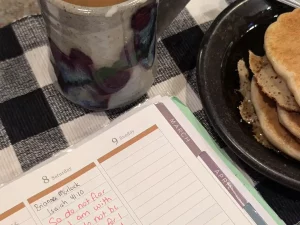 A close-up view of a ceramic mug filled with coffee, a plate with stacked pancakes, and an open planner showing notes for March 8 and 9, including a handwritten Bible verse from Isaiah 41:10. The background features a black and white checkered tablecloth.