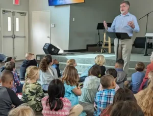 A group of children seated on the carpeted floor of a church or community center, facing a speaker standing near a microphone and music stand. The stage behind the speaker features a stool, microphone stand, and equipment. A screen displays text partially visible at the top, and a door with exit signs is visible in the background.