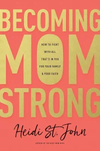 This is the book cover for *Becoming MomStrong* by Heidi St. John. The design features large gold text on a coral-colored background, with the title "Becoming Mom Strong" prominently displayed. The subtitle reads, "How to Fight With All That's in You for Your Family & Your Faith." Below that is the author's name, "Heidi St. John," written in a cursive script, along with "Author of The Busy Mom Blog."