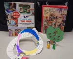 Two children's Bible storybooks, a colorful paper snake, a green paper tree, and two wooden peg dolls on display.