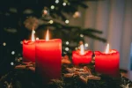 Four red candles lit on an evergreen Advent wreath with wooden stars and festive ornaments, set against a softly blurred Christmas tree and warm holiday lights.