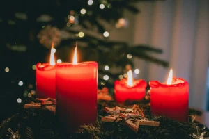 Four red candles lit on an evergreen Advent wreath with wooden stars and festive ornaments, set against a softly blurred Christmas tree and warm holiday lights.