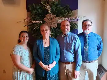 Four Lanse Free Church staff members standing indoors in front of a large floral arrangement with greenery, purple flowers, and a decorative bow.