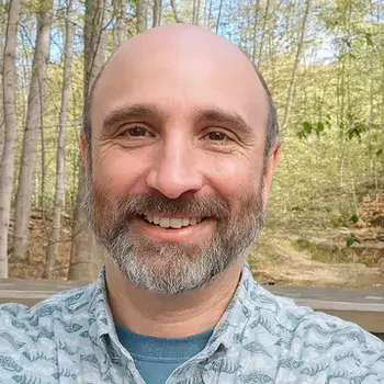 Pastor Matt Mitchell smiles at the camera while standing outdoors in a wooded area. He is wearing a light-colored shirt with a dinosaur pattern. Sunlight filters through tall trees in the background, creating a peaceful forest setting.