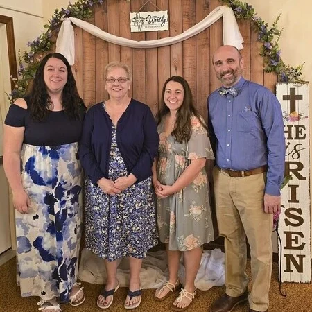 Four staff members of Lanse Free Church stand side by side in front of a decorative wooden backdrop with floral garland, white drapery, and signs reading “Worthy Is the Lamb” and “HE IS RISEN.” From left to right: Jenni English (Administrative Assistant), Cindy Green (Custodian), Jordyn Skacel (Director of Family Ministries), and Pastor Matt Mitchell. All are dressed in semi-formal attire, smiling in a warm indoor setting.